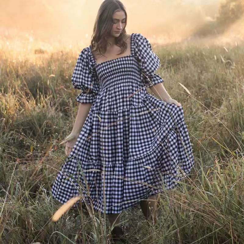 Front view of the Pixi Dress in black and white gingham pattern showcasing square neckline, voluminous puff sleeves, smocked bodice, and flowing tiered midi skirt. Dress photographed in a sunlit meadow