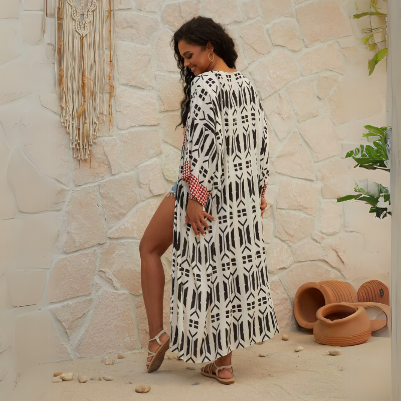 Curly brunette model smiles wearing Ethola Kimono from Shop The Kei Swim collection with denim shorts and tan sandals. Bohemian ankle-length beach cover-up kimono with an earthy geometric print and waist tie. Lightweight, flowy, and perfect for vacations.
