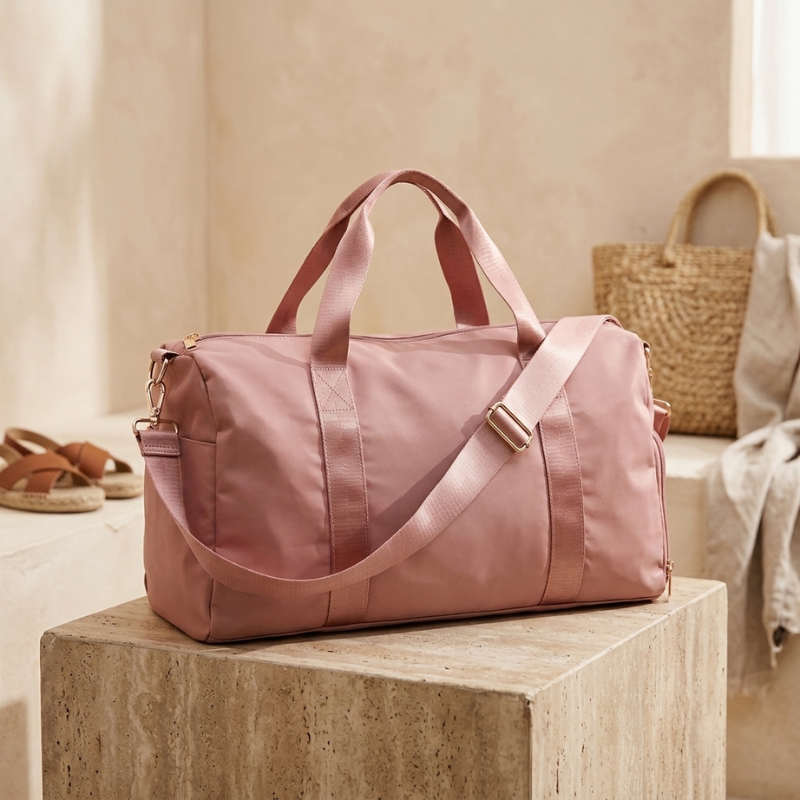A dusty pink nylon duffel bag with dual top handles and a matching adjustable shoulder strap, resting on a textured stone block in a warm, neutral-toned interior setting.
