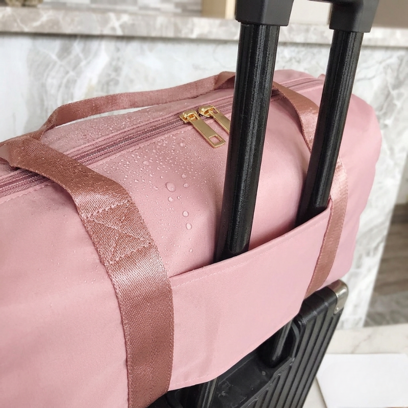 A dusty pink waterproof duffel bag with rose gold satin straps and gold zipper pulls, shown with its black rolling suitcase handle inserted into the side pocket. Water droplets are visible on the surface of the bag, demonstrating its water-resistant feature, set against a marble background.