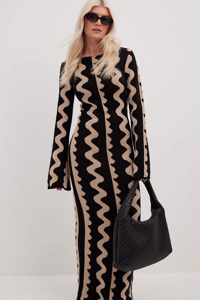 Model wears Gemini Knit Dress by ShoptheKei with a black leather tote bag and oval shaped black sunglasses. Sweater dress maxi length with long flare sleeves. Body conscious. Mod 1960's style dress with vertical wavy squiggle line patterns.