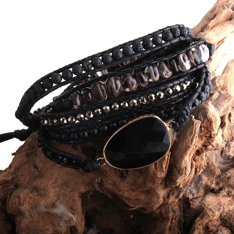 Deep, Volcanic black and earthy tone Bohemian style wrap bracelet with a large center stone and several colorful smaller stones set in a natural cording. The bracelet rests on driftwood. Boho Crystal Wrap Bracelet at Shop the Kei