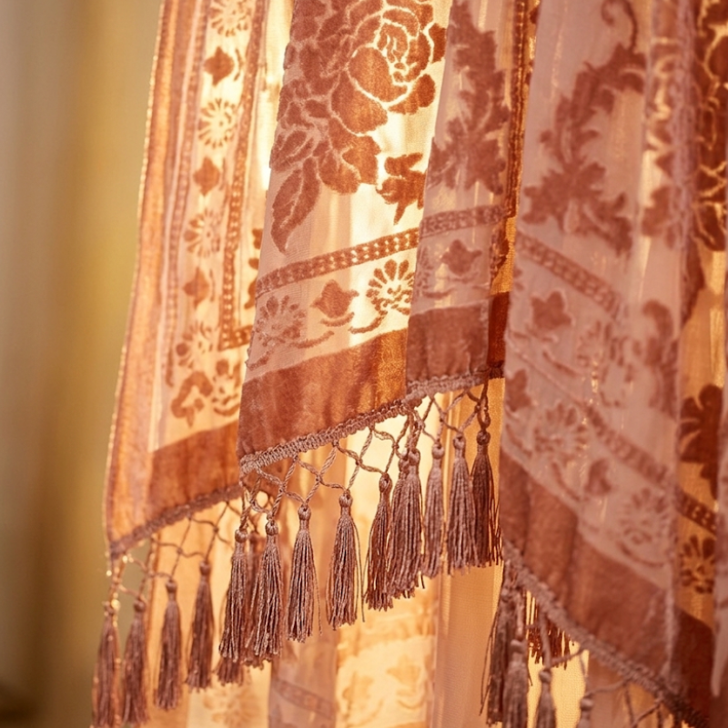Backlit close-up of the Kindle Shawl's sheer floral burnout velvet pattern and tassel hem glowing against window light.