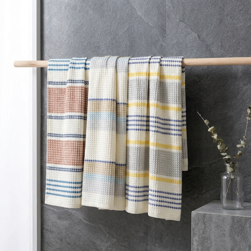 The PetalDry Striped Cotton Bath Towel in Earthy, Blue Breeze, and Lemonade hanging together from a wooden rod against a slate stone wall, with a eucalyptus stem in a glass bottle nearby.