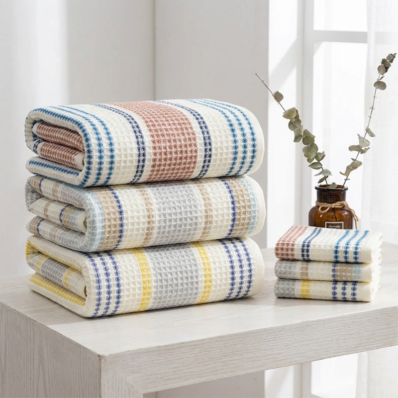 A stack of large PetalDry Bath Towels in Earthy, Blue Breeze, and Lemonade folded beside a matching stack of smaller sizes, showing the range of towel sizes available, styled with a eucalyptus stem on a sunlit wood table.