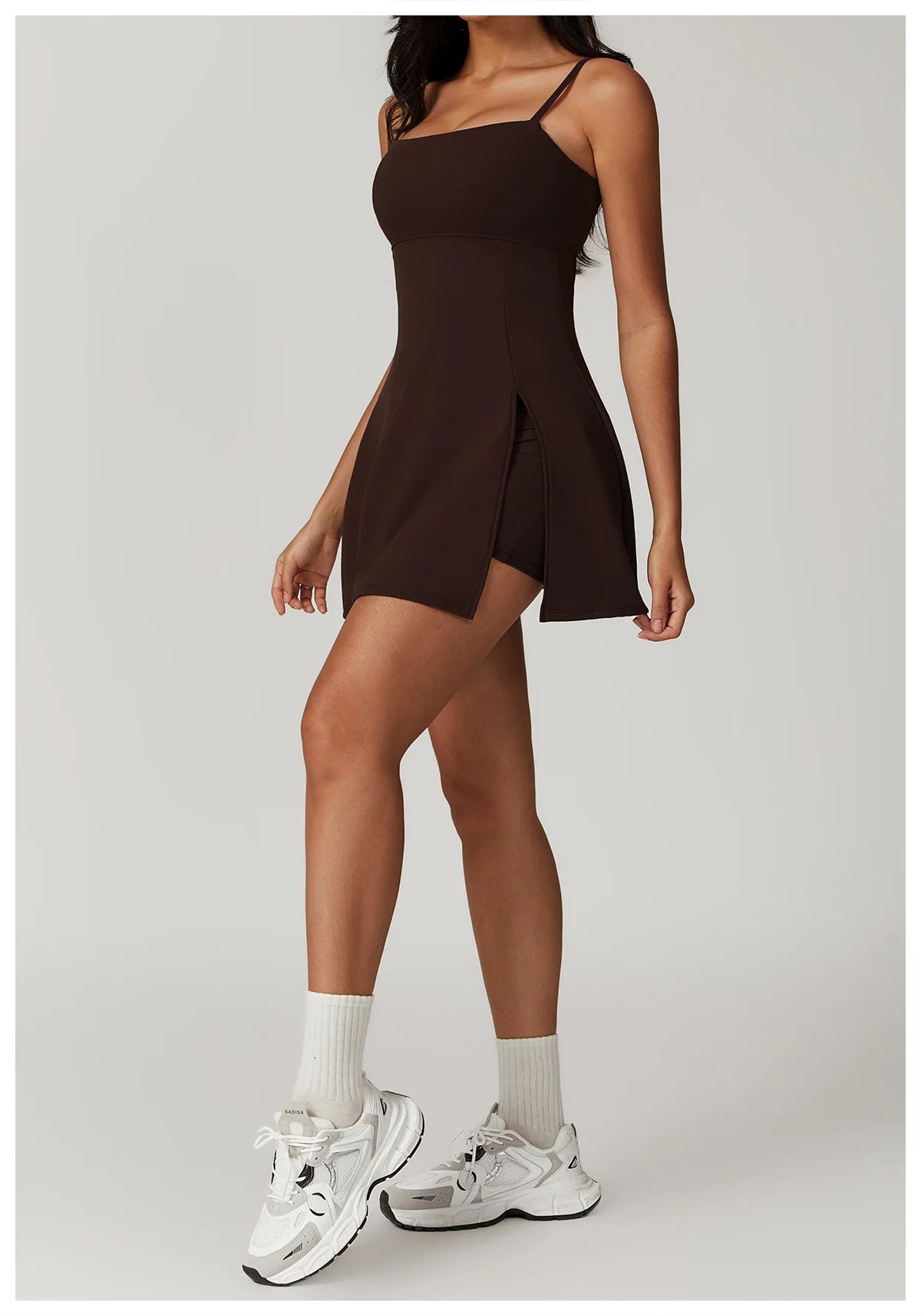Front view of the Bree Yoga Dress in "Espresso" color. A mini dress with thin spaghetti straps, straight neckline, fitted bodice, and short A-line skirt. Model is in motion with white ankle socks and chunky athletic sneakers against a neutral background.