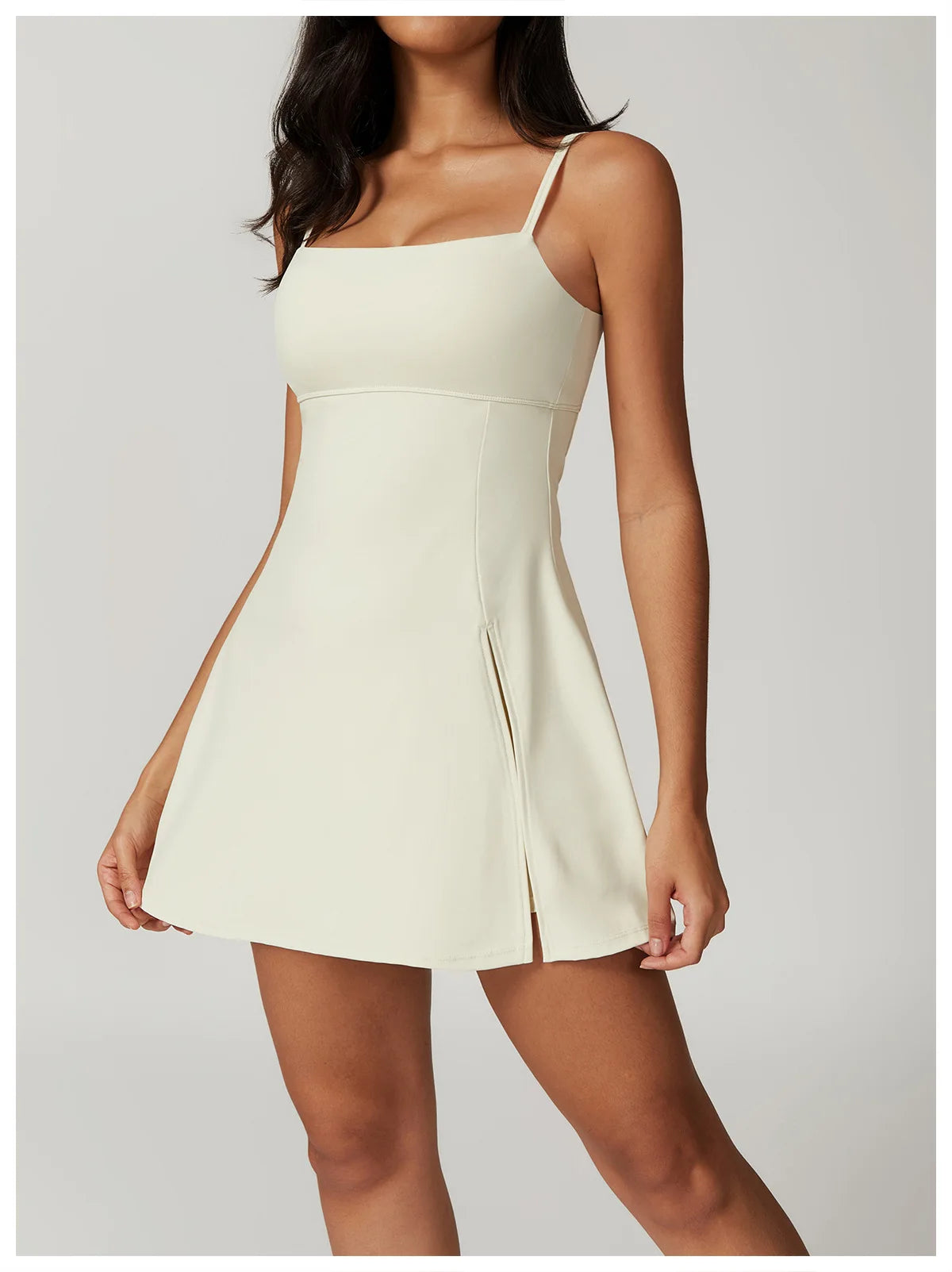 Close up view of the Bree Yoga Dress in creme color. Features thin spaghetti straps, straight neckline, fitted bodice, and A-line skirt with a pronounced side slit. Mini-length dress photographed against a neutral background.