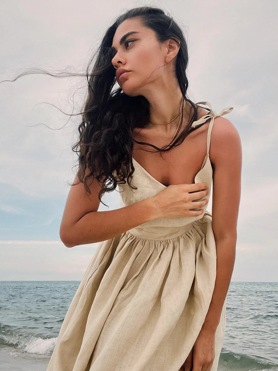 Isla Summer Dress from Shop the Kei - Natural linen dress with gathered waist and tied spaghetti straps, worn by a brunette model in beach waves leaning forward to showcase the natural linen texture and color.