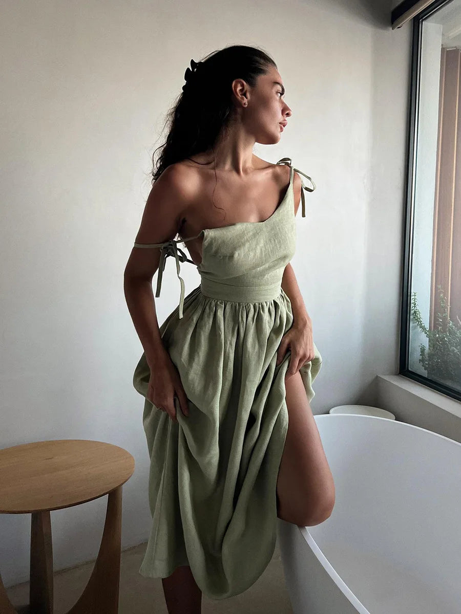 Isla Summer Dress from Shop the Kei - Sage green linen dress with gathered waist, worn by a woman standing near a window with a wooden side table, showcasing elegant and minimalist design