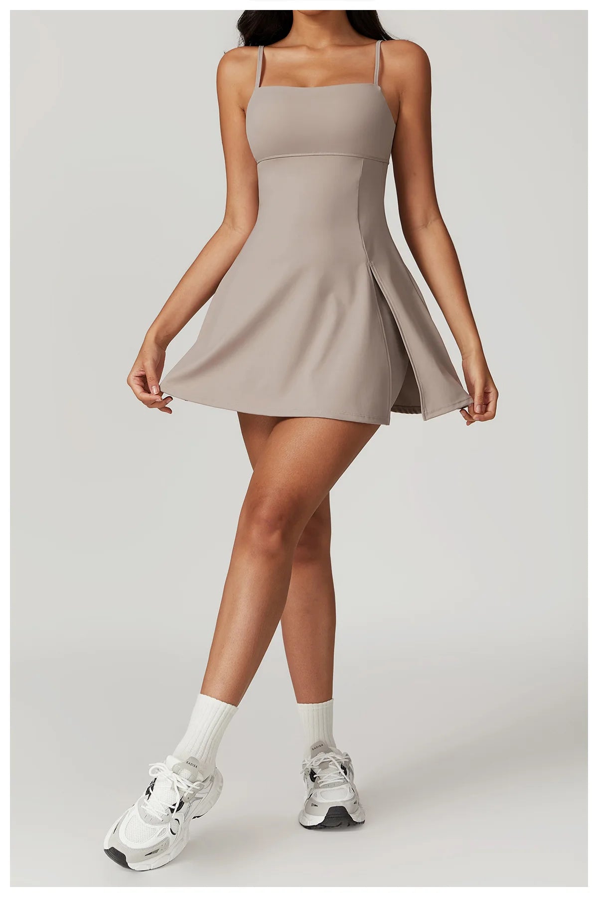 Front view of the Bree Yoga Dress in taupe "Dark Linen" color. A mini dress with thin spaghetti straps, straight neckline, fitted bodice, and short A-line skirt. Model curtsies with white ankle socks and chunky athletic sneakers against a neutral background
