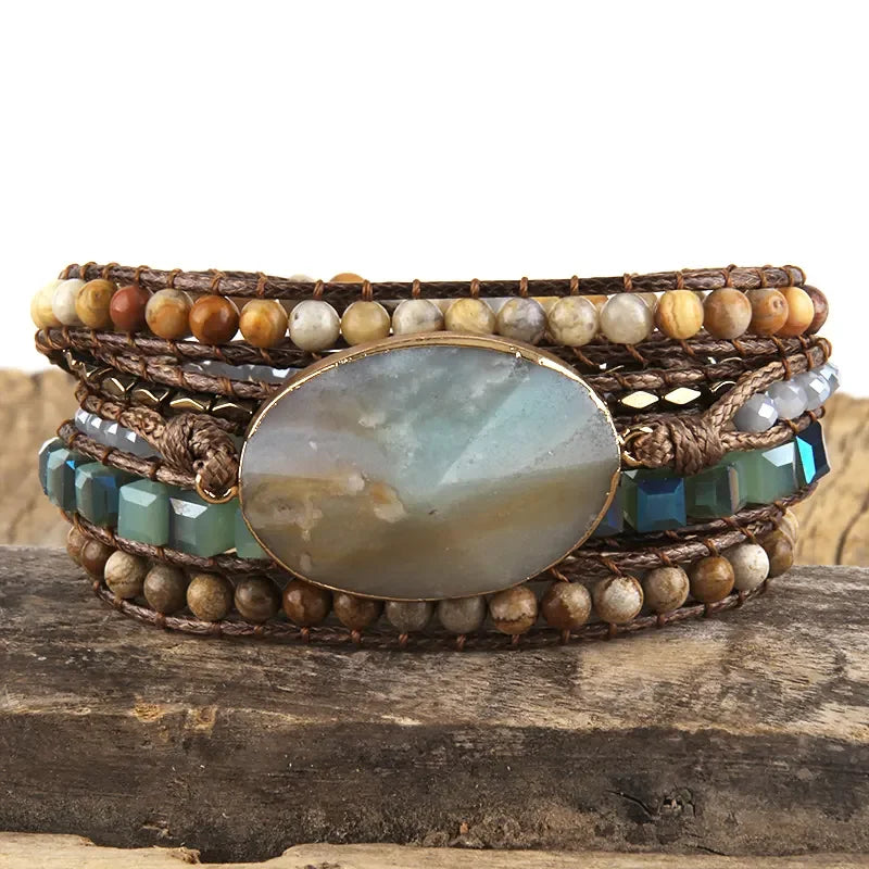 Aquatic greens and warm earthy tone Bohemian style wrap bracelet with a large center stone and several colorful smaller stones set in a natural cording. The bracelet rests on driftwood. Boho Crystal Wrap Bracelet at Shop the Kei