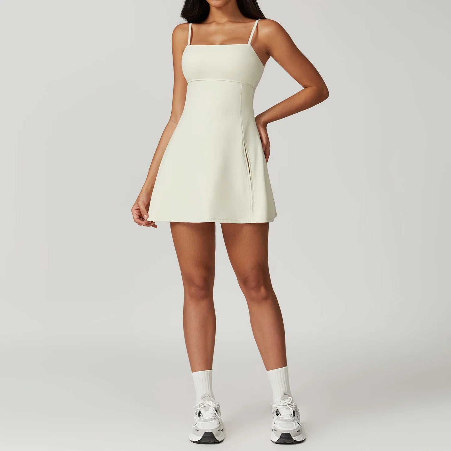 Front view of the Bree Yoga Dress in cream/off-white color. A mini dress with thin spaghetti straps, straight neckline, fitted bodice, and short A-line skirt. Model styled with white ankle socks and chunky athletic sneakers against a neutral background.