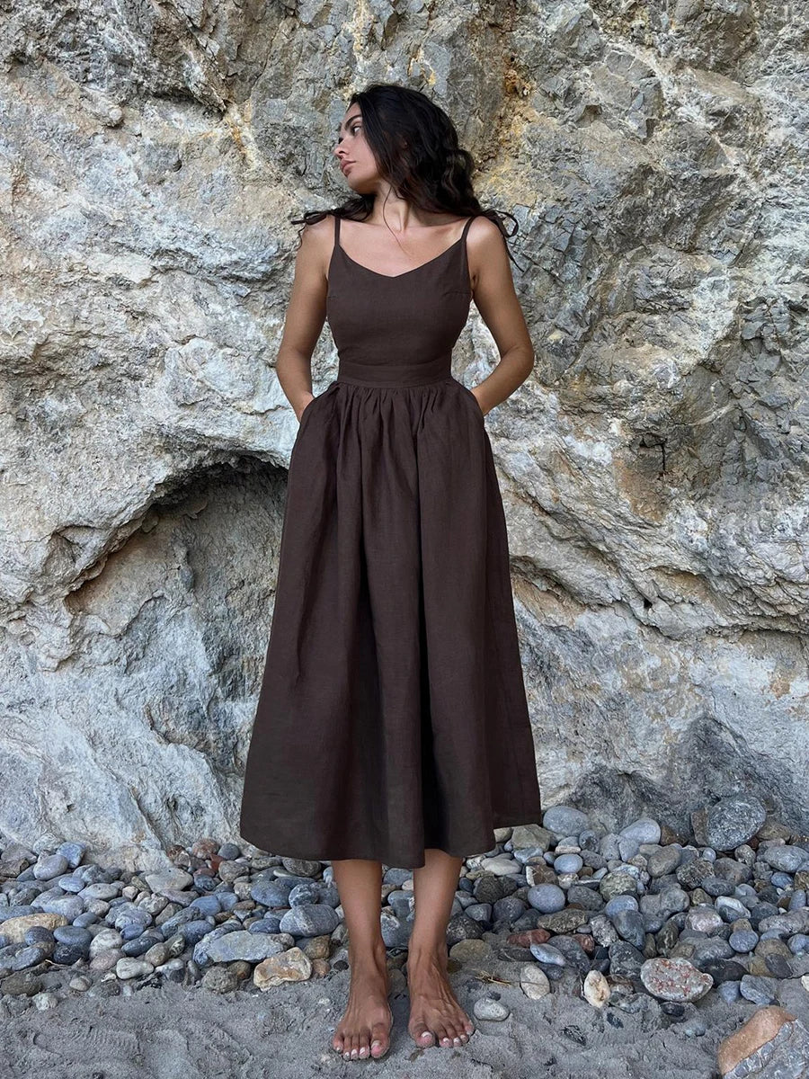 Isla Summer Dress from Shop the Kei -Brunette model wearing a coconut brown linen dress with gathered waist and v neckline and spaghetti straps. Dress fits at the bust and flares at the waist gathers. Photographed against a natural stone background.