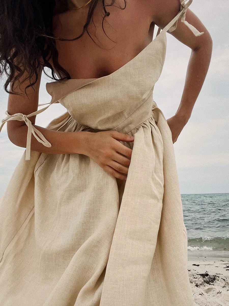 Isla Summer Dress from Shop the Kei -Close up of natural linen dress with gathered waist to show soft textile texture and lining construction. Spaghetti straps elegantly drape off the shoulders of a woman leaning forward with brunette waves softly swept up by a warm breeze on the beach