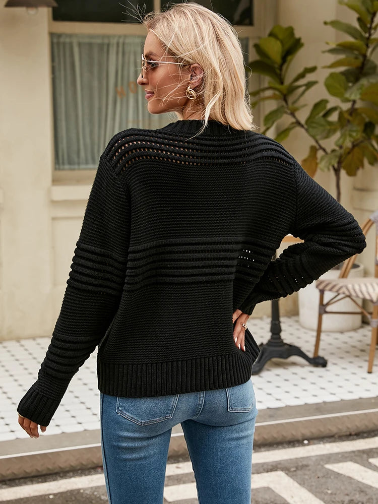 Lea Sweater from Shop the Kei - Back view of a model wearing a relaxed fit black knitted sweater with horizontal openwork pattern detail and long sleeves, paired with light blue jeans. The model has long blonde hair and accessorizes with round sunglasses and gold earrings. The outfit is styled for casual spring or summer wear, photographed outside a cafe with minimal architectural elements.