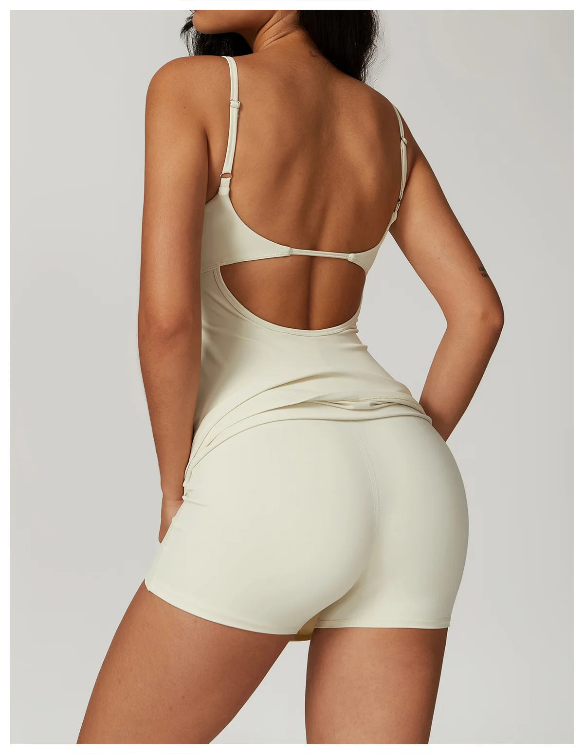 Back view of the Bree Yoga Dress in creme color. Shows the open-back design with thin adjustable straps crossing the back, A-line skirt held up to reveal hidden shorts. Photographed against a neutral background.