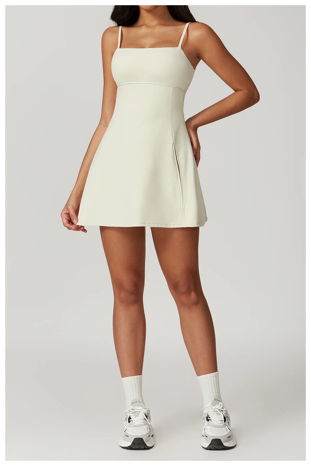 Front view of the Bree Yoga Dress in creme color. A mini dress with thin spaghetti straps, straight neckline, fitted bodice, and short A-line skirt. Model poses with white ankle socks and chunky athletic sneakers against a neutral background.