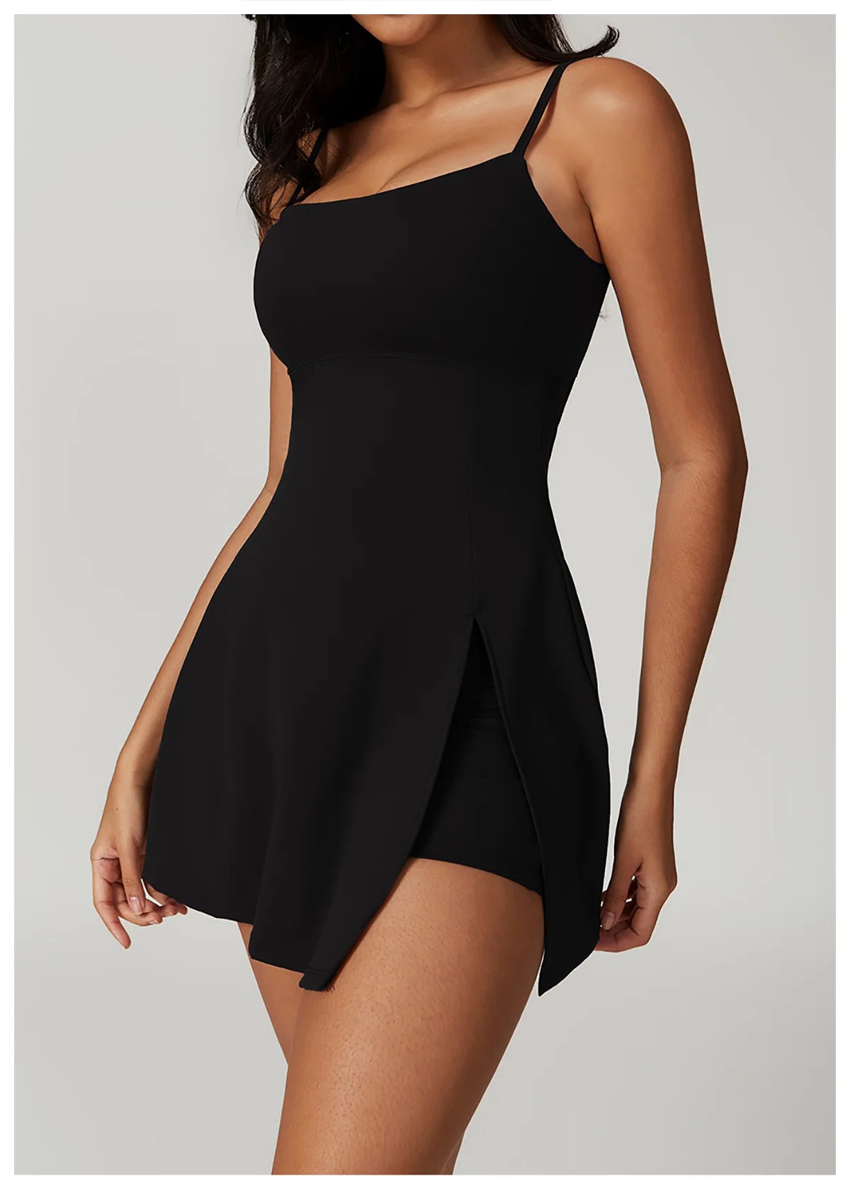 Close up view of the Bree Yoga Dress in black color. Features thin spaghetti straps, straight neckline, fitted bodice, and A-line skirt with a pronounced side slit. Mini-length dress photographed against a neutral background.