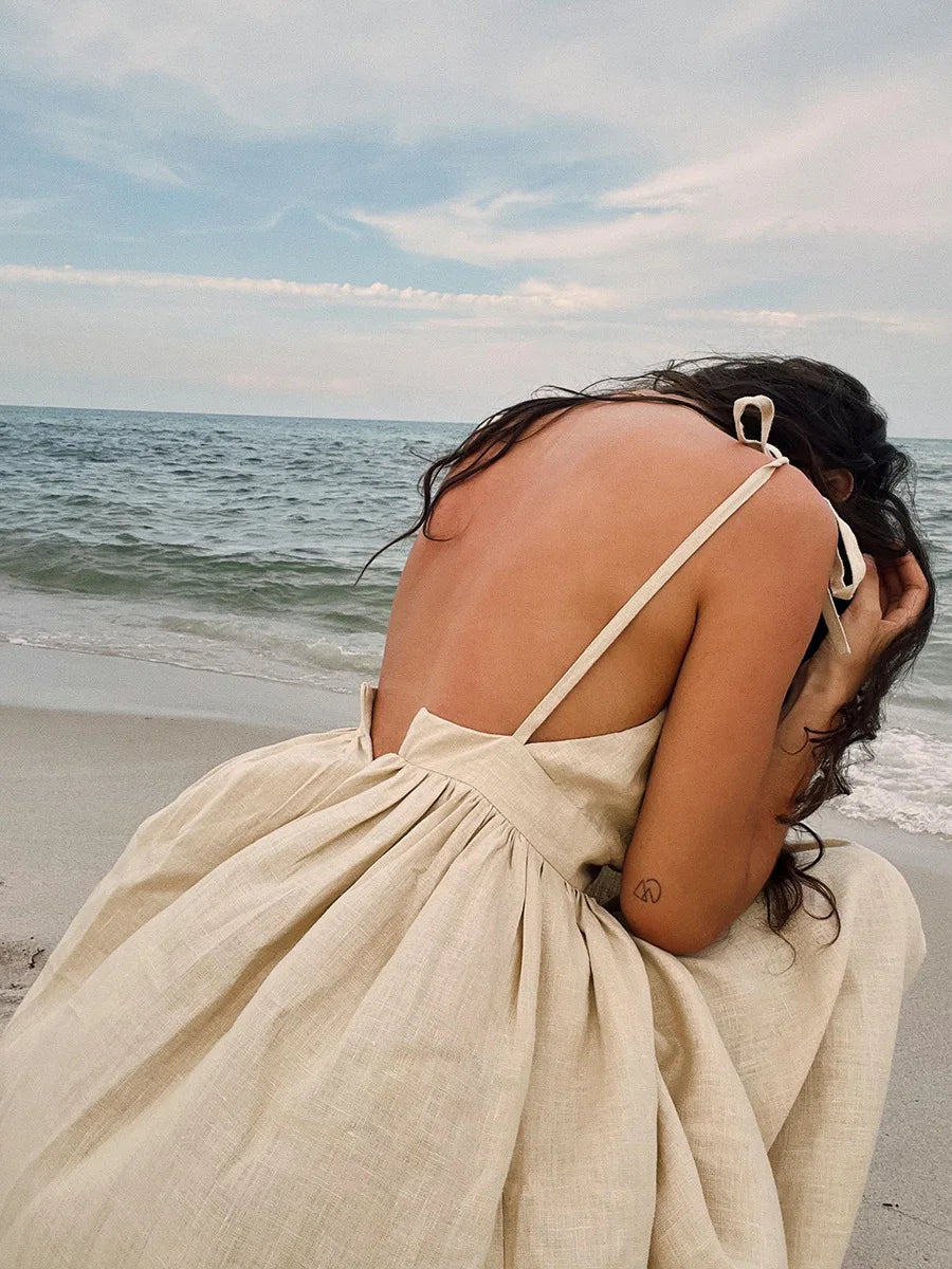 Isla Summer Dress from Shop the Kei -Close up back view of natural linen dress with gathered waist to show soft textile texture and fullness. Spaghetti straps are tied and worn by a model with brunette waves softly swept up by a warm breeze on the beach