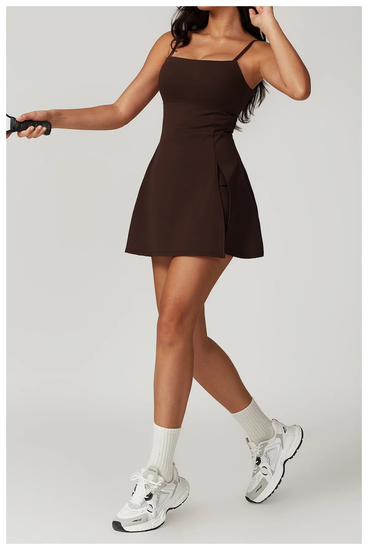 Front view of the Bree Yoga Dress in "Espresso" color. A mini dress with thin spaghetti straps, straight neckline, fitted bodice, and short A-line skirt. Model is in motion with white ankle socks and chunky athletic sneakers against a neutral background.