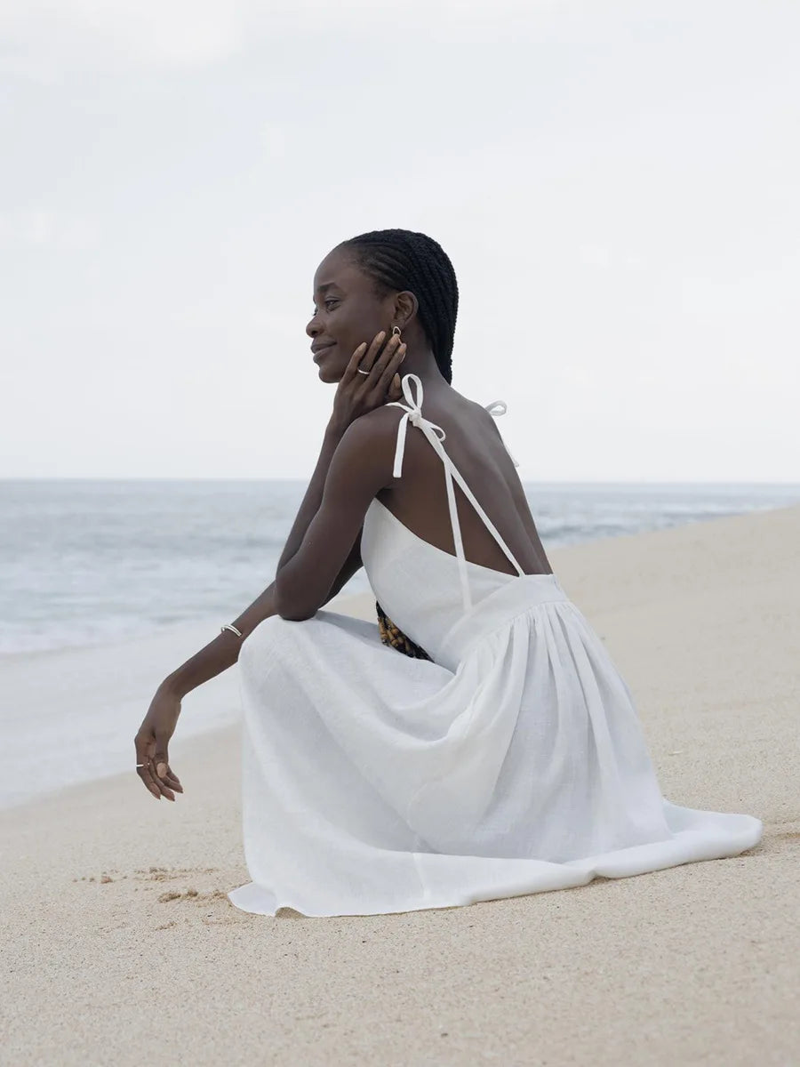 Isla Summer Dress from Shop the Kei - Soft white linen dress with gathered waist, worn by a woman sitting on the sand gazing blissfully at the shore. Paired with minimalist jewelry and long beaded braids
