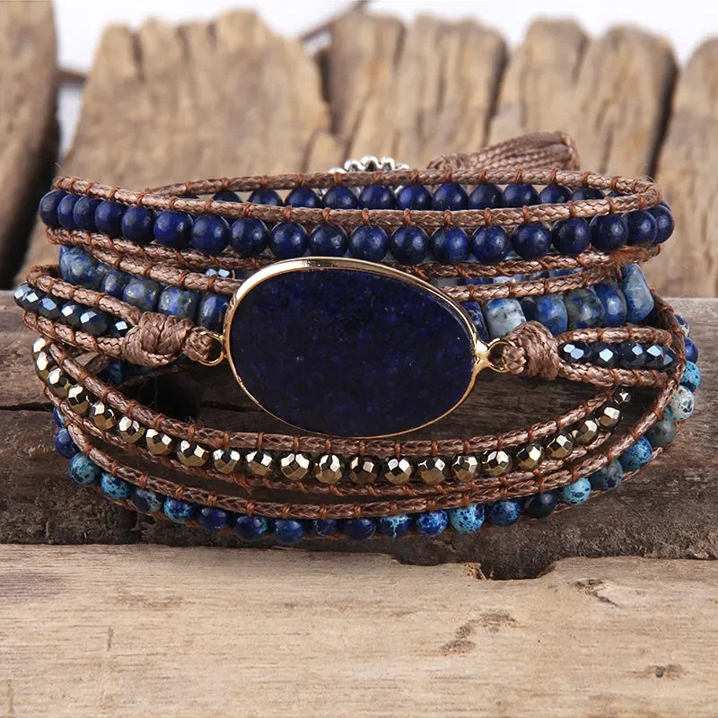 Navy blues and earthy tone Bohemian style wrap bracelet with a large center stone and several colorful smaller stones set in a natural cording. The bracelet rests on driftwood. Boho Crystal Wrap Bracelet at Shop the Kei