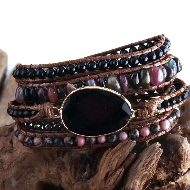 Black, purple and earthy tone Bohemian style wrap bracelet with a large center stone and several colorful smaller stones set in a natural cording. The bracelet rests on driftwood. Boho Crystal Wrap Bracelet at Shop the Kei