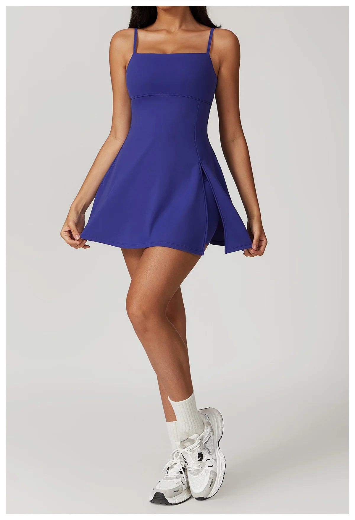 Front view of the Bree Yoga Dress in deep bright "Blue Glass" color. A mini dress with thin spaghetti straps, straight neckline, fitted bodice, and short A-line skirt. Model styled with white ankle socks and chunky athletic sneakers against a neutral background.
