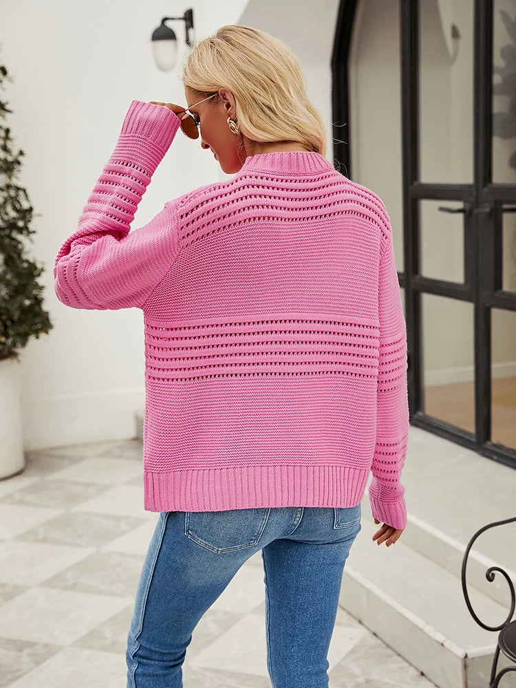 Lea Sweater from Shop the Kei - Back view of a model wearing a relaxed fit bright pink knitted sweater with horizontal openwork pattern detail and long sleeves, paired with light blue jeans. The model has long blonde hair and accessorizes with round sunglasses and gold earrings. The outfit is styled for casual spring or summer wear, photographed outside a modern building with minimal architectural elements.