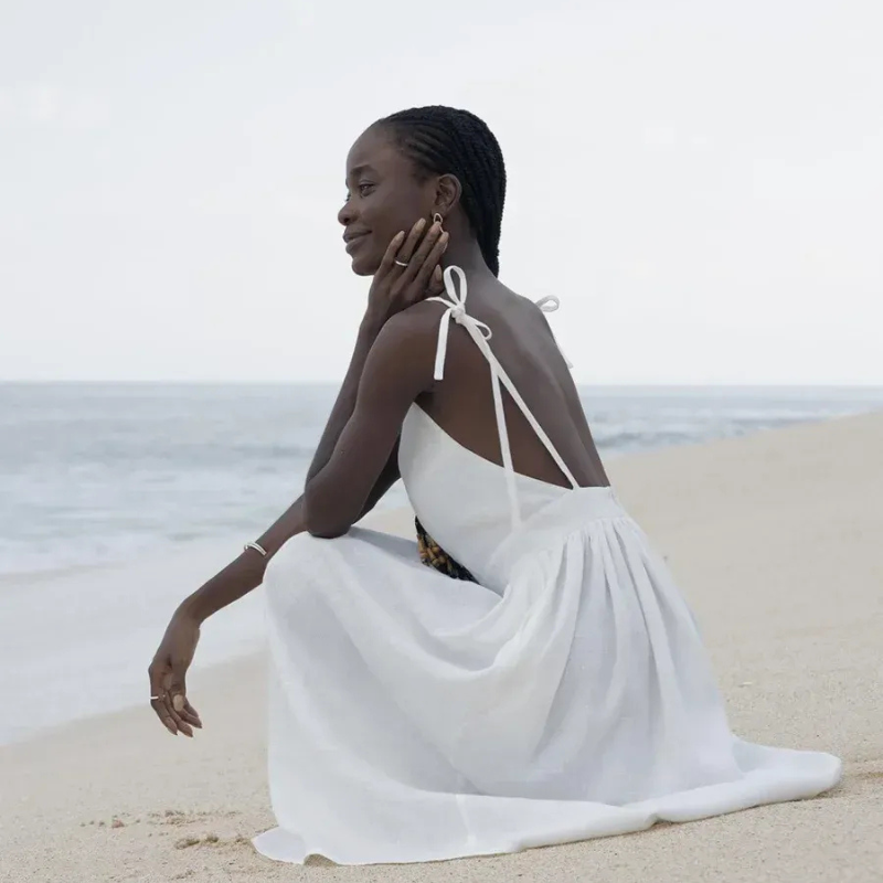 Isla Summer Dress from Shop the Kei - Soft white linen dress with gathered waist, worn by a woman sitting on the sand gazing peacefully at the shore. Paired with minimalist jewelry and long beaded braids
