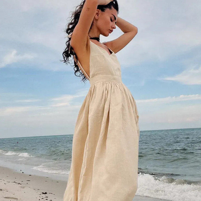 Isla Summer Dress from Shop the Kei - Natural linen dress with gathered waist, worn by a woman standing on the sand gazing peacefully at the shore.