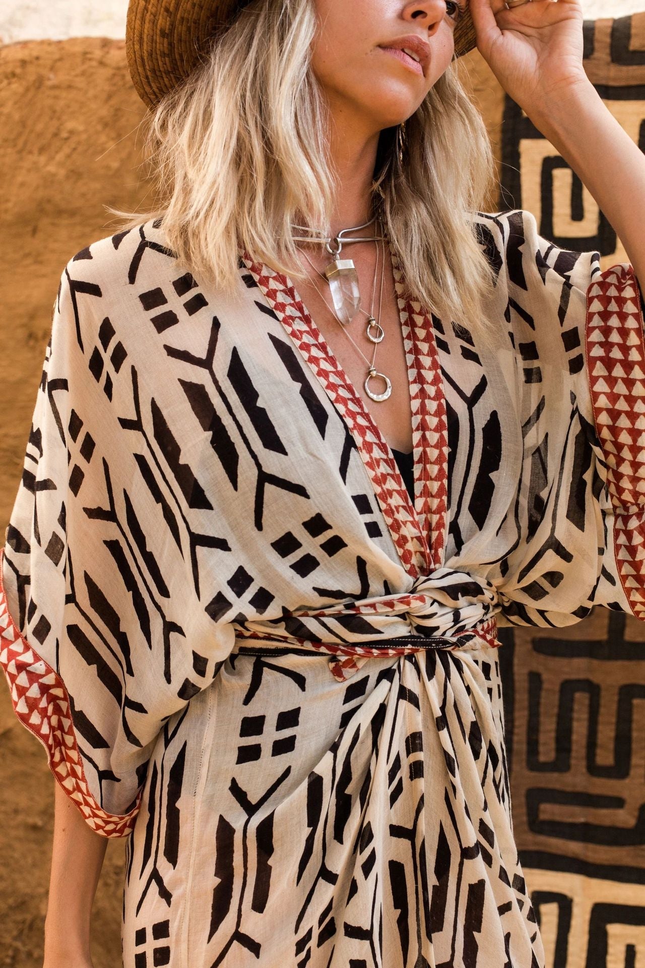 Close up of blonde model wearing Ethola Kimono from Shop The Kei Swim collection with a sun hat. Image depicts earthy geometric print, wrapped waist tie, and draped fabric. Model pairs the kimono with sterling silver boho jewelry