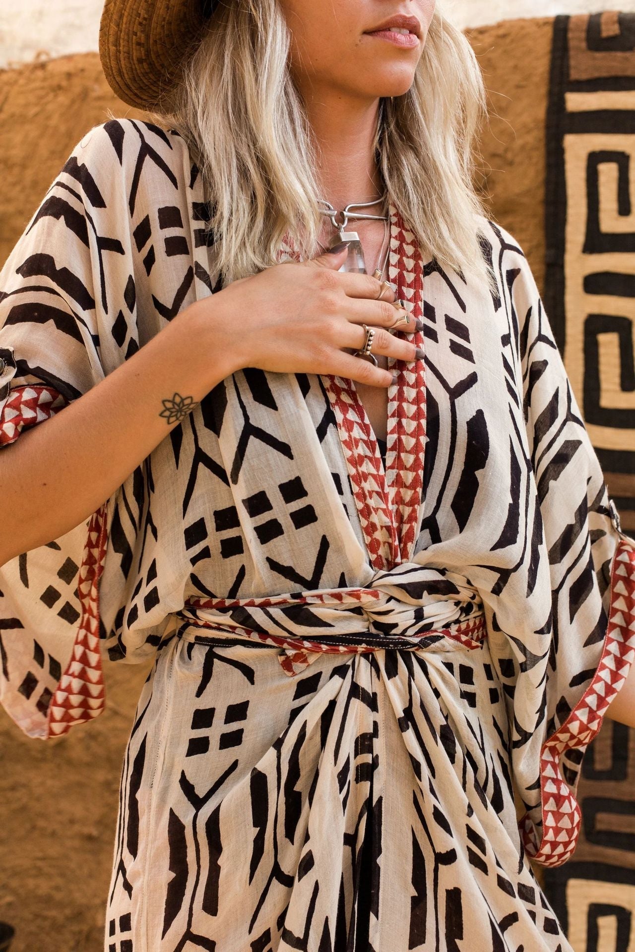 Close up of blonde model wearing Ethola Kimono from Shop The Kei Swim collection with a sun hat. Image depicts earthy geometric print, wrapped waist tie, and draped fabric. Model pairs the kimono with sterling silver boho jewelry