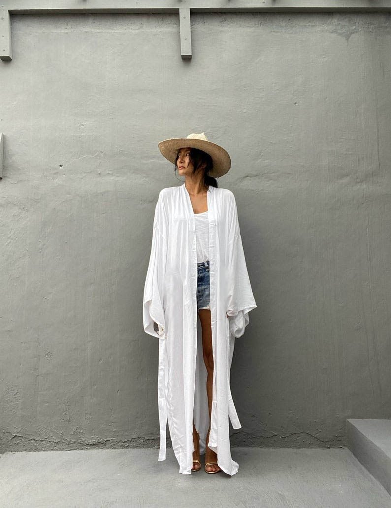 Front view of model wearing the white Sole Kimono from Shop the Kei with a white tank, denim shorts, straw hat, beach tote and gold heels. A Bohemian-style ankle-length beach cover-up kimono with wide sleeves and matching waist sash, perfect for vacation, poolside, or post-swim lounging.