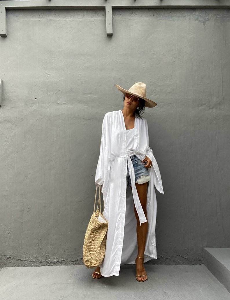 Front view of model wearing the white Sole Kimono from Shop the Kei with a white tank, denim shorts, straw hat, beach tote and gold heels. A Bohemian-style ankle-length beach cover-up kimono with wide sleeves and matching waist sash, perfect for vacation, poolside, or post-swim lounging.