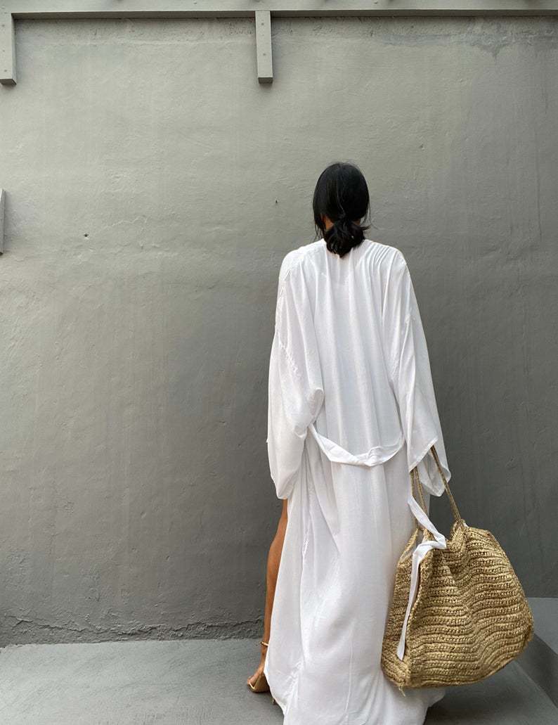 Back view of model wearing the white Sole Kimono from Shop the Kei with a beach tote. A Bohemian-style ankle-length beach cover-up kimono with wide sleeves and matching waist sash, perfect for vacation, poolside, or post-swim lounging.