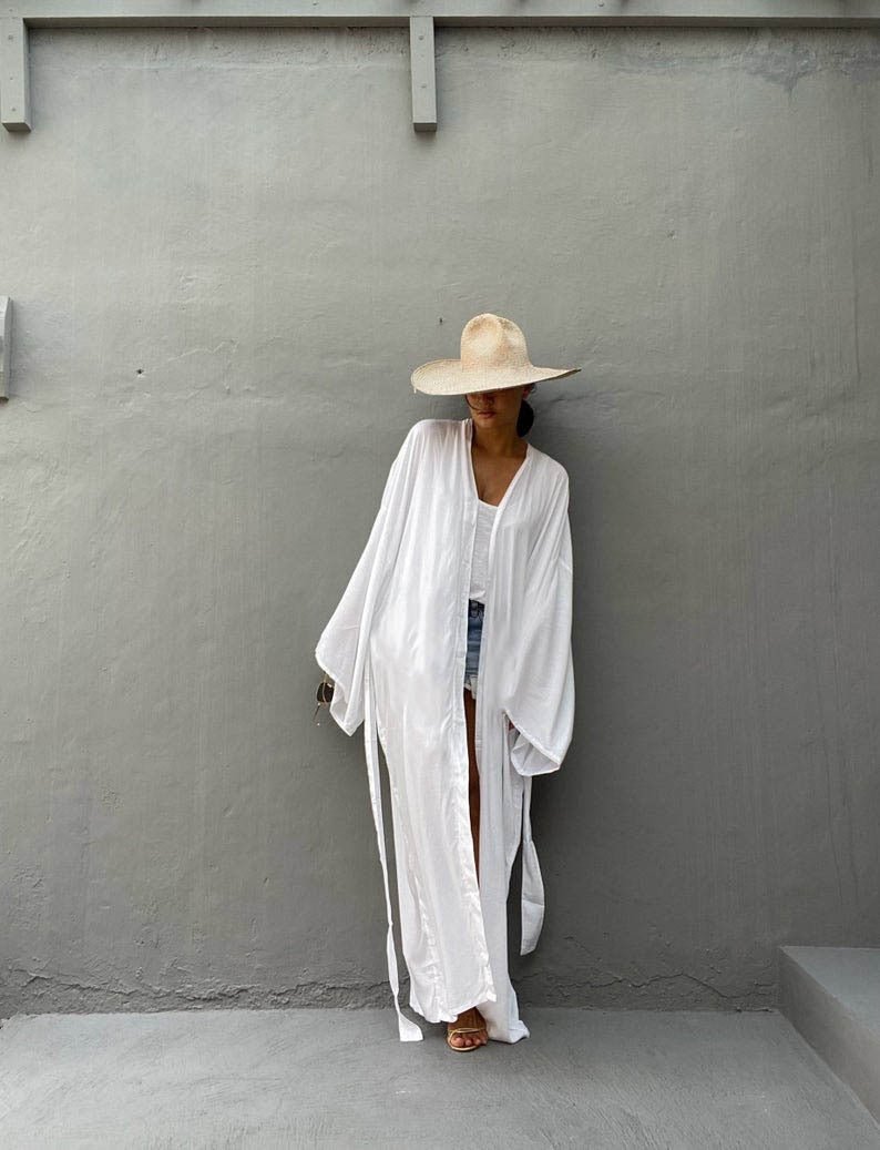 Front view of model wearing the white Sole Kimono from Shop the Kei with a white tank, denim shorts, straw hat, beach tote and gold heels. A Bohemian-style ankle-length beach cover-up kimono with wide sleeves and matching waist sash, perfect for vacation, poolside, or post-swim lounging.