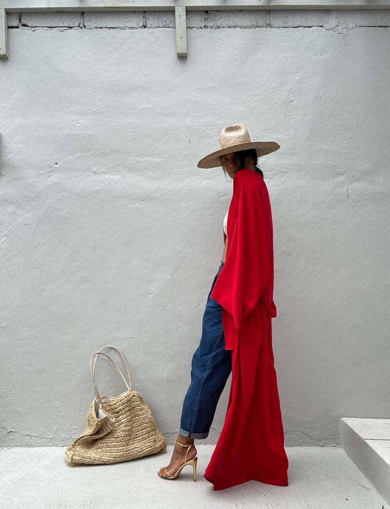 Side view of model wearing the red Sole Kimono from Shop the Kei shown with a white bikini top, baggy jeans, gold heels, straw hat and beach tote . A Bohemian-style ankle-length beach cover-up kimono with wide sleeves and matching waist sash, perfect for vacation, poolside, or post-swim lounging.