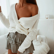 Close-up front view of a model wearing the Aries Sweater in milk white featuring a distinctive twisted front knot design. The chunky knit sweater is styled with a ruffled gray mini skirt, worn off-shoulder with a wrap-style closure that creates a cropped silhouette. Sweater photographed in a white comfortable living room. Showcasing the sweater's chunky rib knit texture, soft yarns, relaxed fit, and versatile styling.