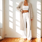 Person wearing a long beige cardigan over a white outfit in a sunlit room.