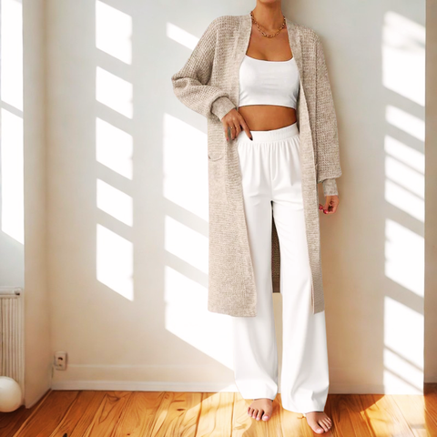 Person wearing a long beige cardigan over a white outfit in a sunlit room.