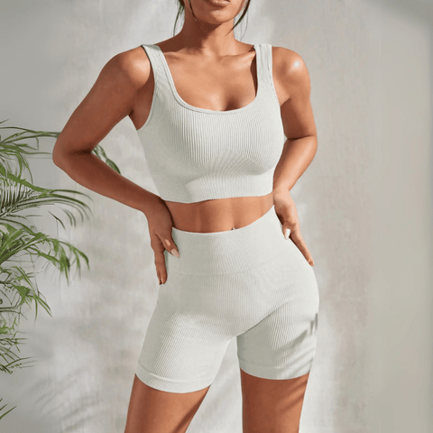 Model wearing Zen Yoga Shorts Set in white. Soft textured rib knit tank top with matching slim fit high waist shorts provide support and comfort during your movements. Model stands with hands on her hips in front of a white background.