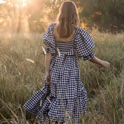 Back view of the Pixi Dress in black and white gingham check pattern, photographed in a sunlit meadow during golden hour. Model walking through tall grass showing the dress's square neckline back design, puff sleeves, fitted bodice, and flowing tiered skirt silhouette against a natural landscape backdrop.