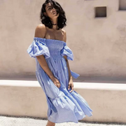 Pixi Dress in light blue gingham pattern featuring off-shoulder design with puff sleeves, smocked bodice, and tiered midi-length skirt. Model shown in front of a neutral beige wall with natural lighting highlighting the dress's airy silhouette.