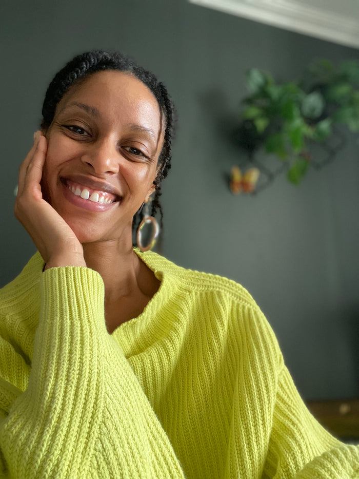 A smiling Kei with dark skin and braided hair rests her head on her hand, wearing a chunky lime green ribbed knit sweater and silver hoop earrings against a dark gray wall.