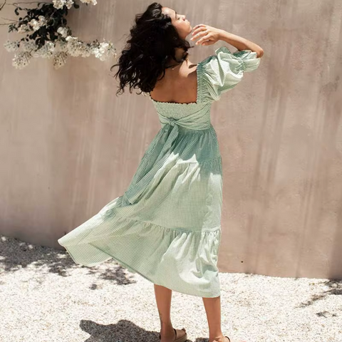 Back view of the Pixi Dress in mint green gingham pattern, featuring a square neckline, puff sleeves, fitted smocked bodice, and tiered midi-length skirt. Model shown in natural lighting against a neutral wall with flowering branches.