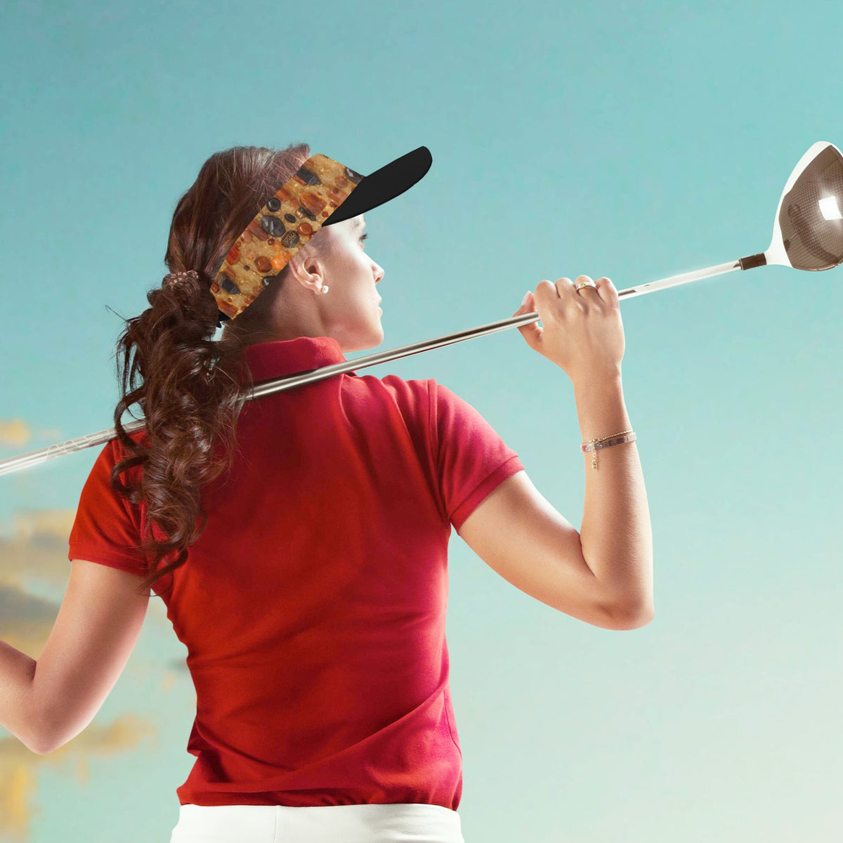 A woman on a golf course wearing a red polo shirt, white pants, and a patterned visor, captured from behind as she swings a driver. The background is a soft blue sky gradient.