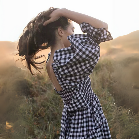 Side view of the Pixi Dress in black and white gingham pattern, featuring a model with her hand in her hair captured during golden hour in a natural field setting. The dress shows puff sleeves, smocked bodice, and tiered midi skirt design, with warm sunset light creating an atmospheric glow.