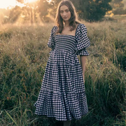 Front view of the Pixi Dress in black and white gingham pattern showcasing square neckline, voluminous puff sleeves, smocked bodice, and flowing tiered midi skirt. Dress photographed in a sunlit meadow