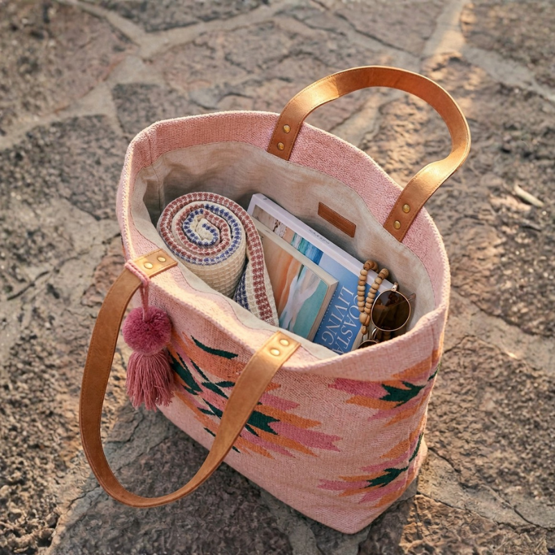 A Blue Breeze PetalDry Bath Towel rolled and tucked inside an open Aztec-print tote bag alongside a magazine, sunglasses, and a beaded bracelet, showing how easily it packs for the beach.